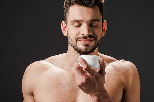 positive naked man with closed eyes holding plastic container with face cream isolated on grey