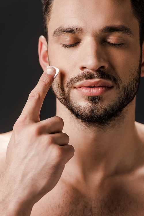 naked man with closed eyes applying face cream isolated on black