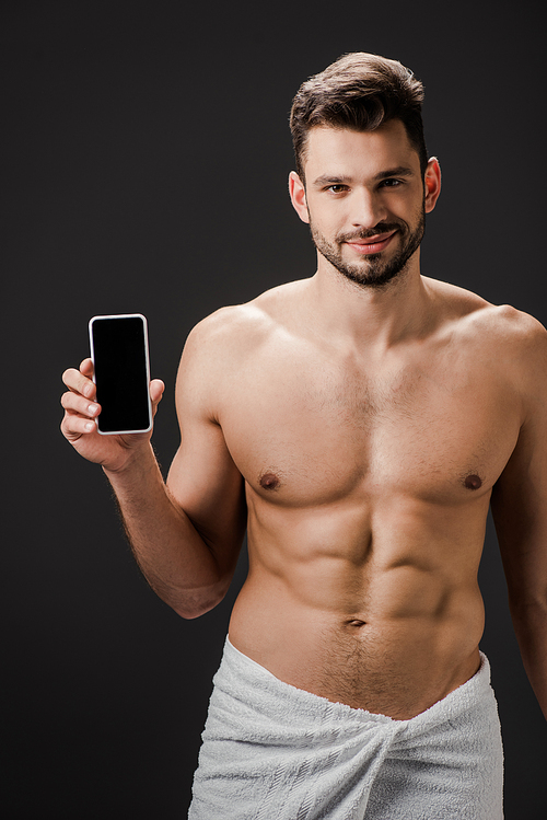 smiling sexy man in towel showing smartphone with blank screen isolated on black