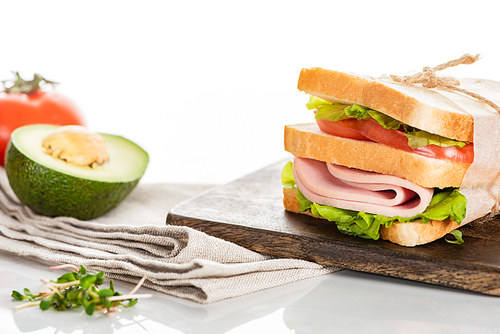 fresh delicious sandwich with sliced sausage and lettuce on wooden cutting board near napkin and avocado on white surface
