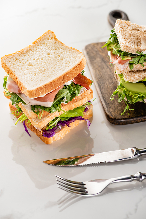 fresh sandwiches with meat on marble white surface with cutlery and wooden cutting board