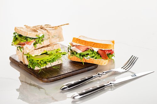fresh sandwiches on wooden cutting board near cutlery on white marble surface