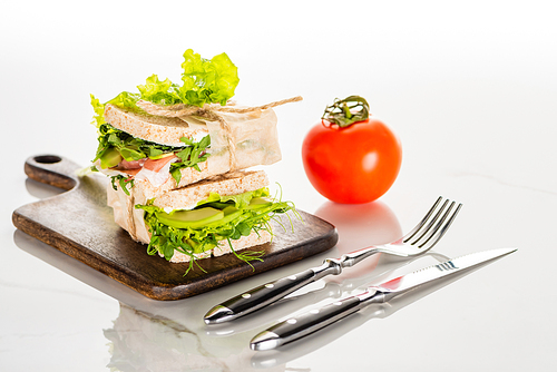 fresh green sandwiches on wooden cutting board near cutlery and tomato on white marble surface