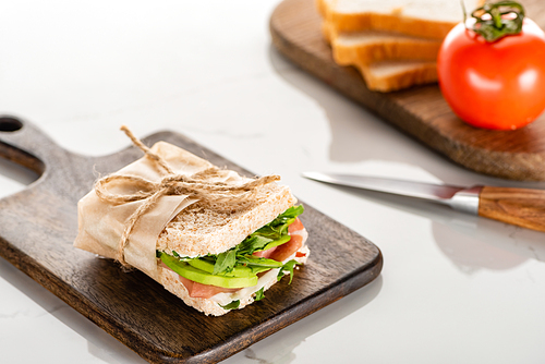 selective focus of fresh green sandwich with avocado and prosciutto on wooden cutting board on white marble surface