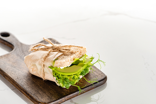 fresh green vegan sandwich with avocado on wooden cutting board on white marble surface