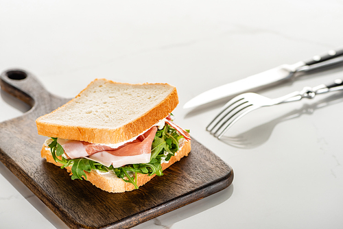 selective focus of fresh sandwich with arugula and prosciutto on wooden cutting board near cutlery on white marble surface