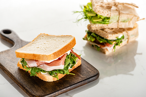 selective focus of fresh green sandwich with prosciutto on wooden cutting board on white marble surface