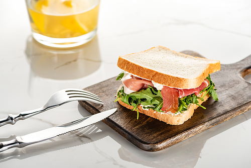 fresh green sandwich with arugula and prosciutto on wooden cutting board with cutlery on white marble surface
