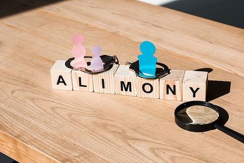 wooden blocks with alimony lettering near family paper cut, magnifier and handcuffs on surface