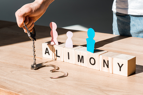 cropped view of man holding handcuffs near wooden blocks with alimony lettering and paper people