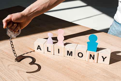 cropped view of man holding handcuffs near wooden blocks with alimony lettering and paper people on desk