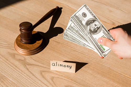 cropped view of man holding dollar banknotes near gavel and paper with alimony lettering on desk