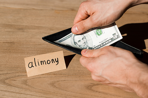 cropped view of man holding wallet and dollar banknote near paper with alimony lettering