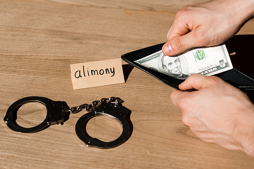 cropped view of man holding wallet and dollar banknote near paper with alimony lettering and handcuffs
