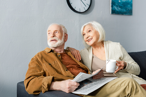 senior man with newspaper and his smiling wife with cup of tea looking away together
