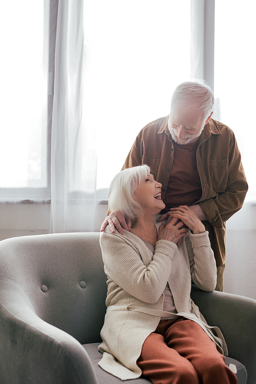 senior man holding hand and touching shoulder of cheerful wife sitting in armchair