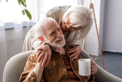 smiling woman hugging happy senior husband sitting with cup of tea