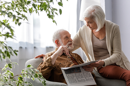 senior woman talking to husband holding newspaper and eyeglasses while sitting in armchair