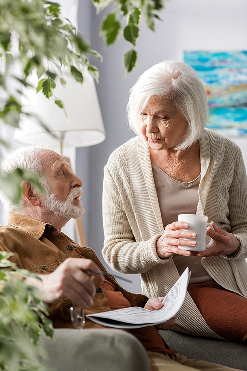 selective focus of senior woman holding cup of tea while talking to surprised husband