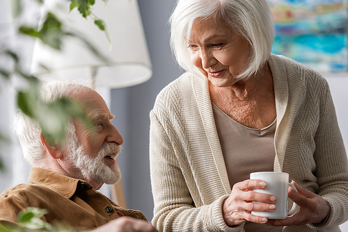 selective focus of smiling senior woman holding cup of tea while talking to husband
