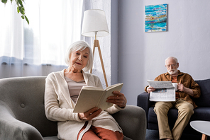 attentive senior woman reading book in armchair near husband with newspaper on sofa