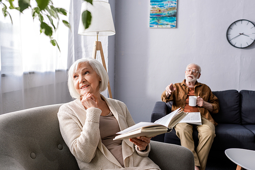 senior man pointing with finger while smiling wife holding book and looking away