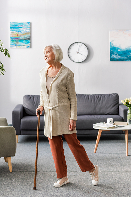 happy senior woman looking away while standing with walking stick at home