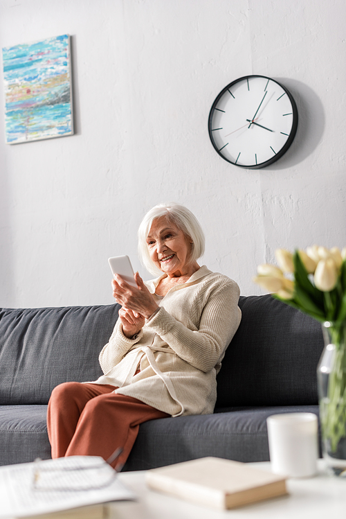 selective focus of happy senior woman using smartphone while sitting on sofa