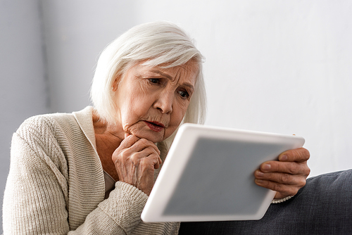 worried senior woman holding hand near chin while using digital tablet
