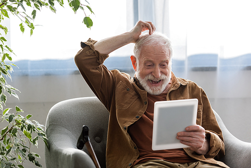 cheerful senior man touching had while using digital tablet in armchair