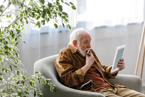 shocked senior man using digital tablet while sitting in armchair