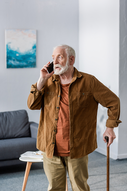 shocked senior man talking on smartphone while standing with walking stick