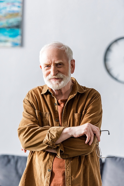 happy senior man smiling at camera while standing with crossed arms and holding eyeglasses