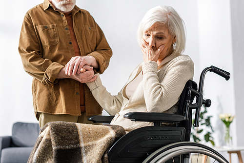 cropped view of man holding hand of disabled wife, sick on dementia, holding hand on face while sitting in .