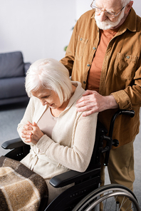 senior man touching shoulder of disabled wife, sick on dementia, sitting in . with bowed head and clenched hands