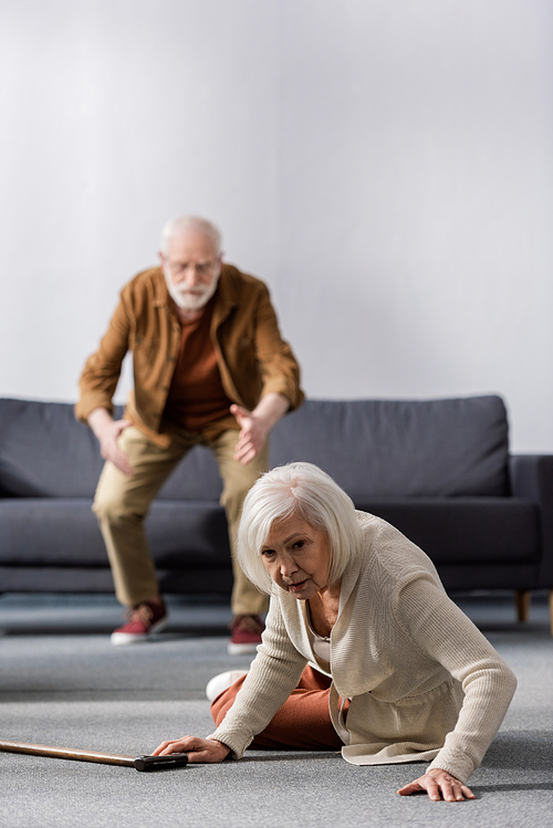 selective focus of senior man hurrying to help wife lying on floor near walking stick