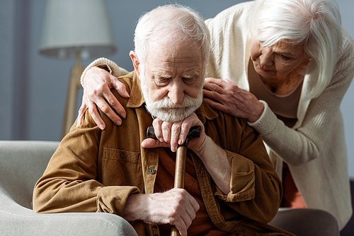 senior man, sick on dementia, sitting with walking stick, while wife looking at him and touching his shoulders