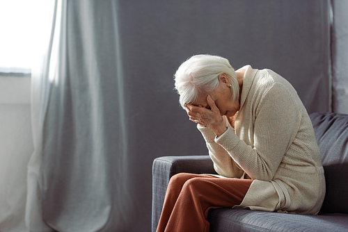 depressed senior woman sitting on sofa with bowed had and obscuring face with hands