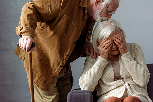 senior man with walking stick kissing head of diseased wife crying and covering eyes with hands