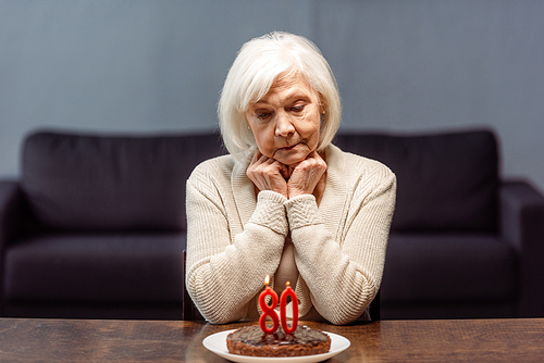 thoughtful, sad senior woman sitting near birthday cake with number eighty and burning candles alone