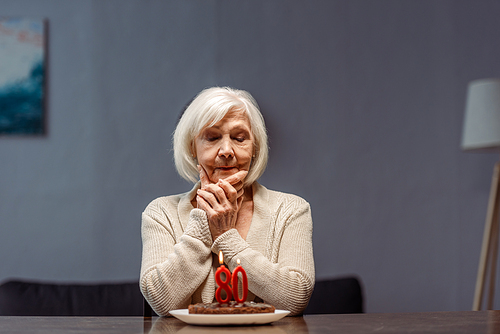 senior, lonely woman touching face while looking at birthday cake with number eighty and burning candles