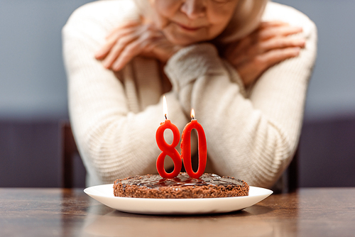 cropped view of senior woman sitting alone near bithday cake with number eighty and burning candles