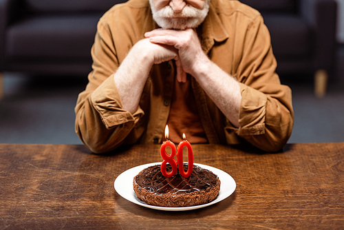 cropped view of lonely senior man leaning on clenched hands and looking at birthday cake with number eighty