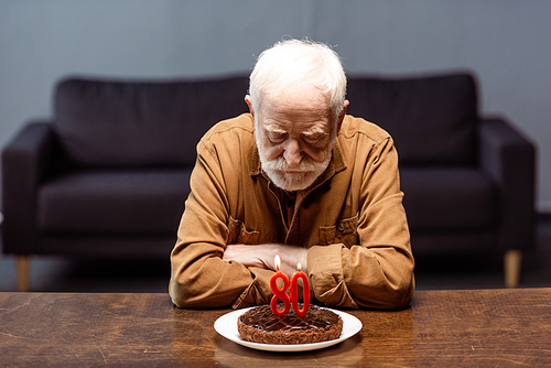 senior lonely man sitting with crossed arms near birthday cake with number eighty