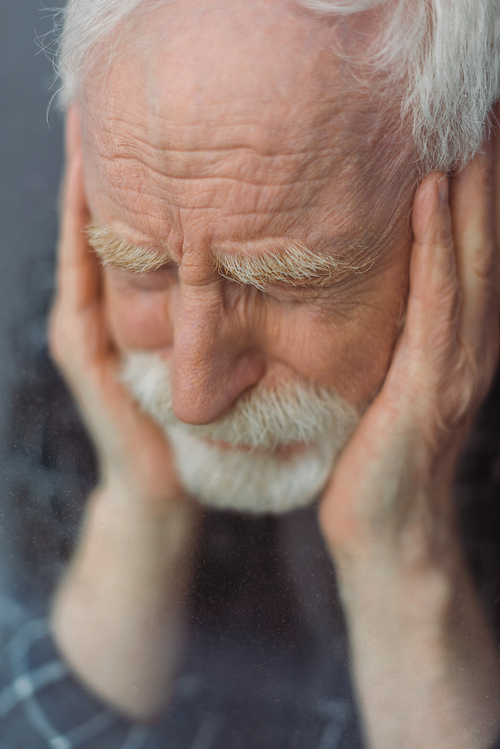 selective focus of senior, depressed man with closed eyes touching face near window glass