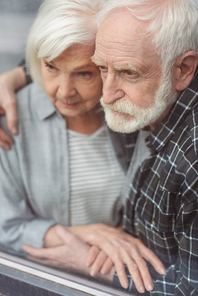 thoughtful senior man and his wife, sick on dementia, holding hands while standing by window