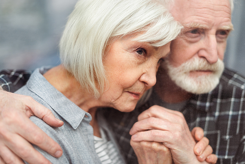 thoughtful senior man holding hands of wife, sick in dementia, while looking through window