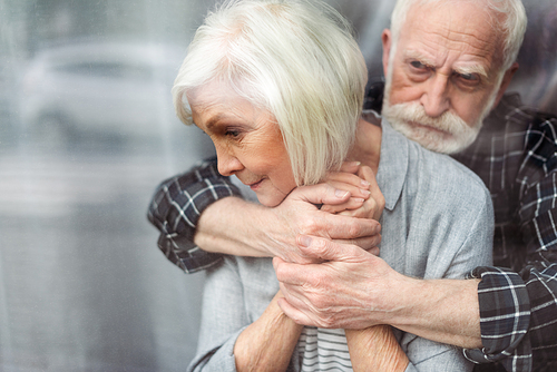 upset senior man hugging wife, sick on dementia, while standing by window