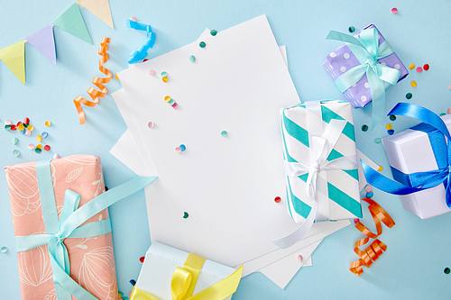 top view of colorful confetti near blank paper and gift boxes on blue background