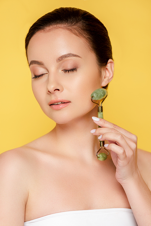 beautiful woman with closed eyes using jade roller on face isolated on yellow